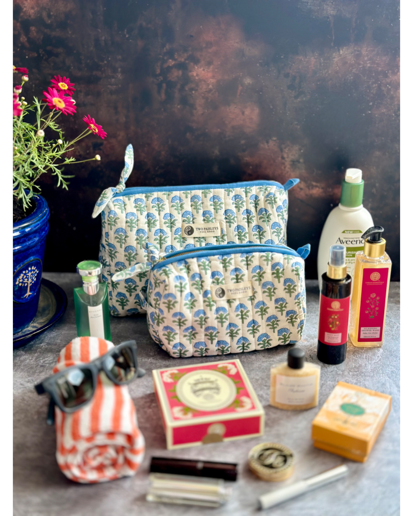 A quilted cosmetic bag with a floral pattern in blue, white, and green shades, displayed with various travel and makeup items like sunglasses, a towel, and bottles.