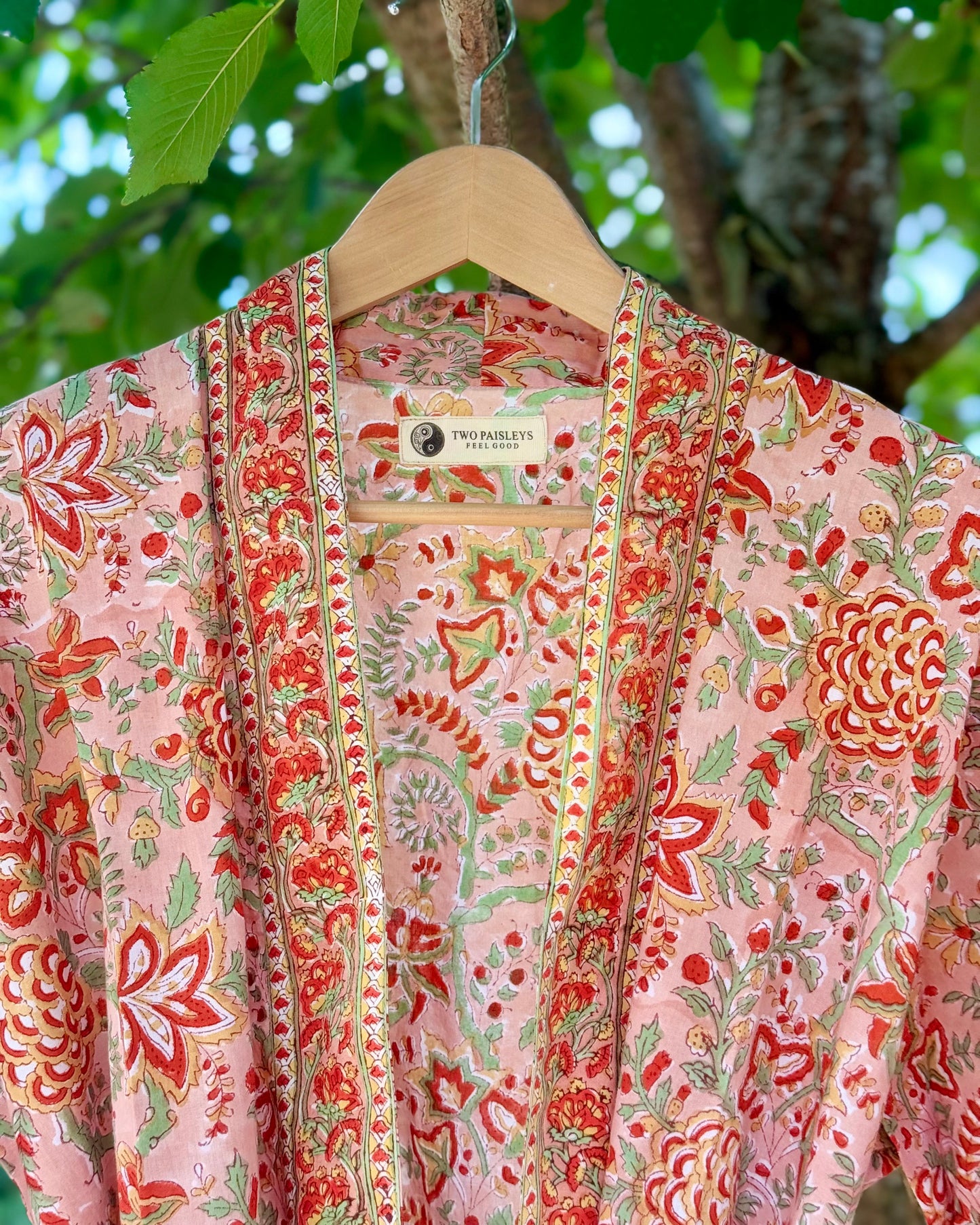 Close-up of a coral pink block printed cotton dressing gown with intricate red and green floral designs and a decorative border, showing the Two Paisleys label.