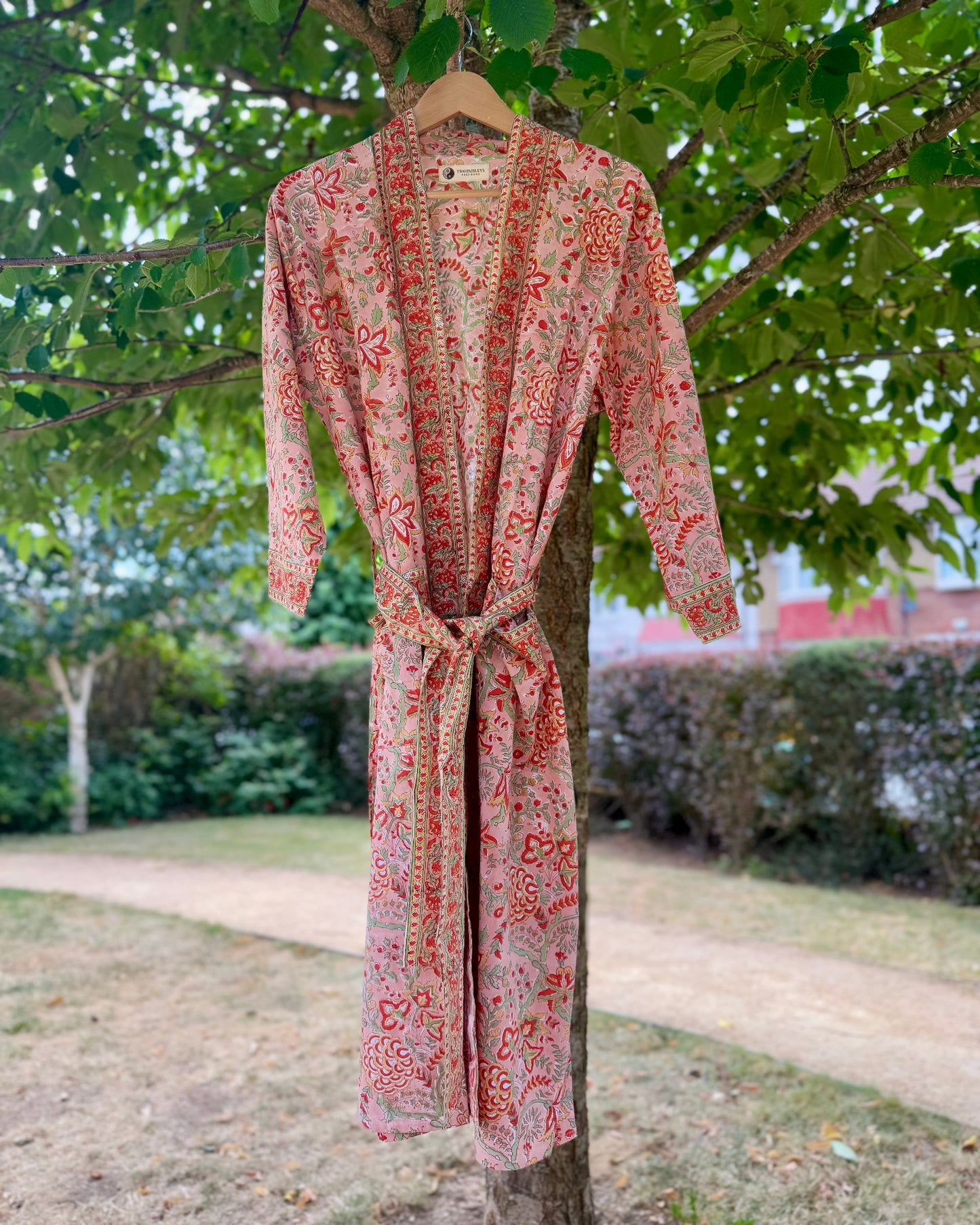 Full front view of a handcrafted coral floral cotton dressing gown with red and green block print, tied at the waist and hanging on a tree outdoors