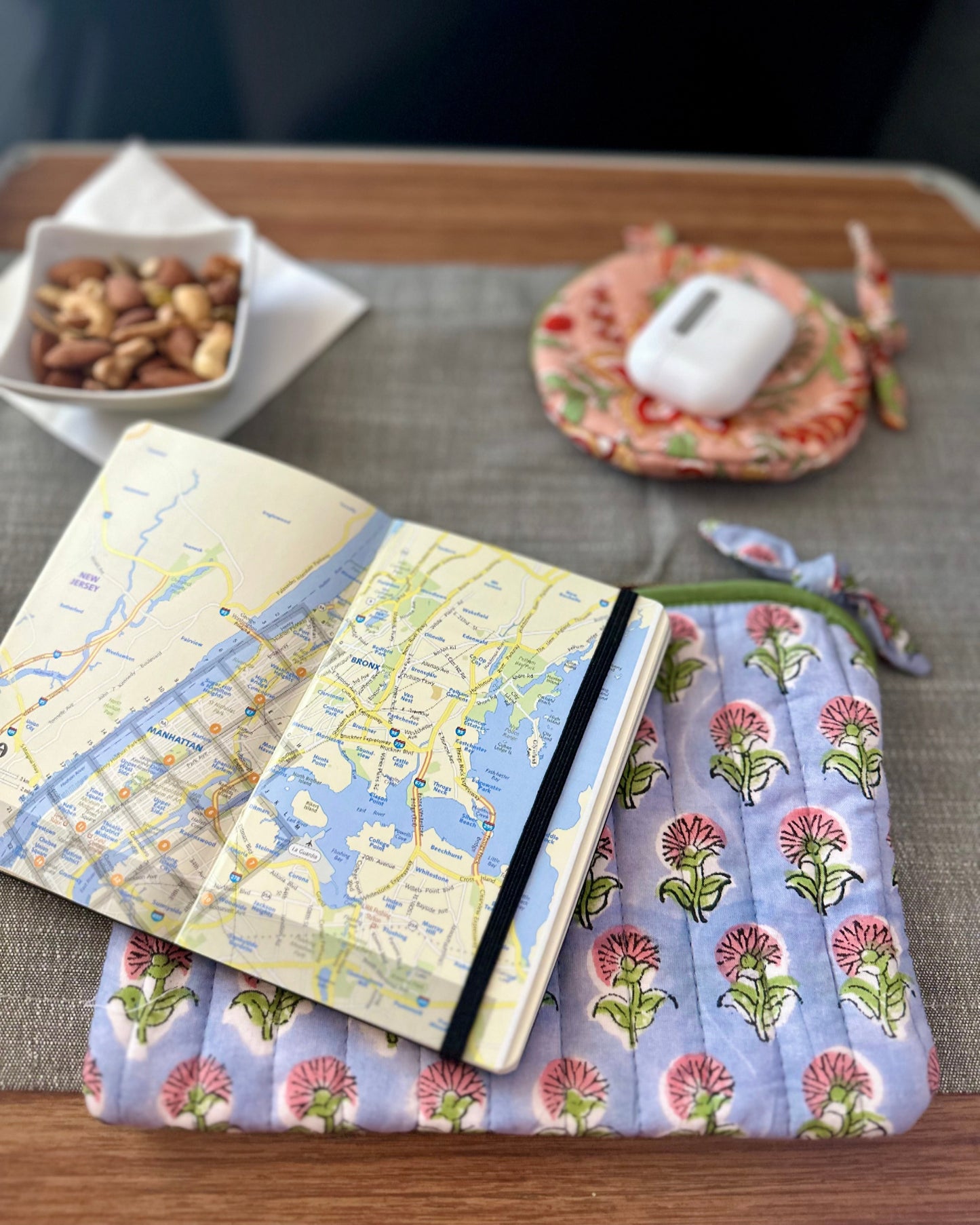 Notebook with map design on a pink & purple floral fabric flat pouch cover for passports, jewellery placed on a table with AirPods or ear buds.
