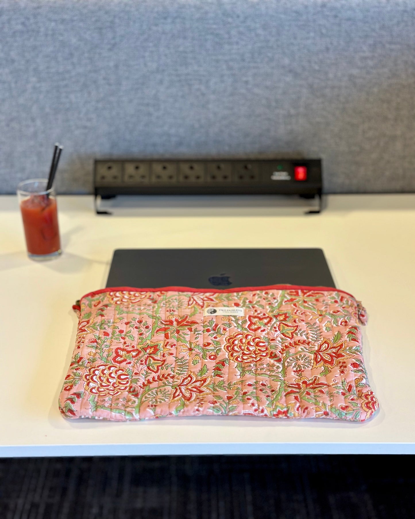 Floral-patterned pouch on a desk with a glass of juice and power strip in the background.