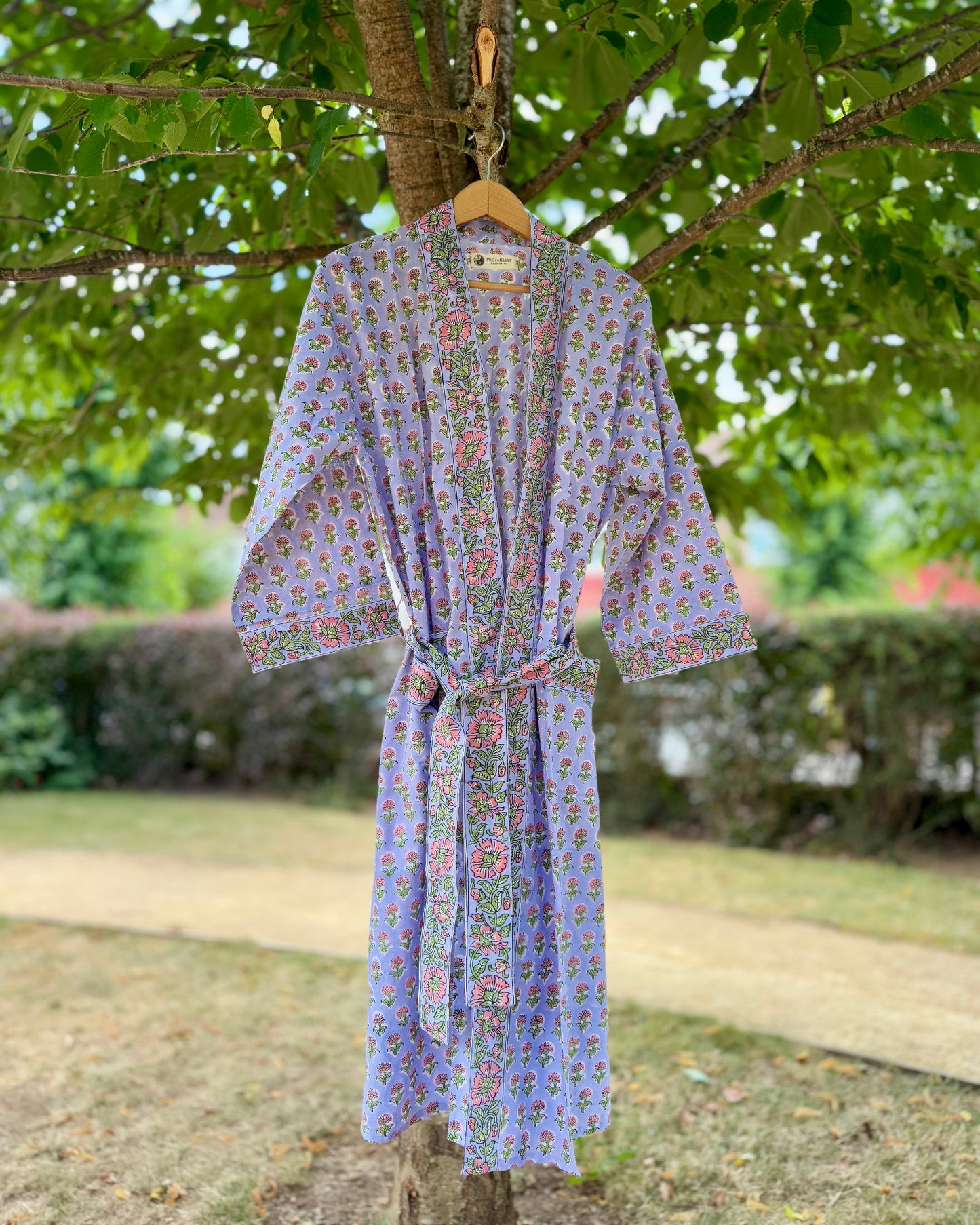 Front view of a floral block printed cotton robe in lilac with pink and green detailing, displayed outdoors on a tree hanger.