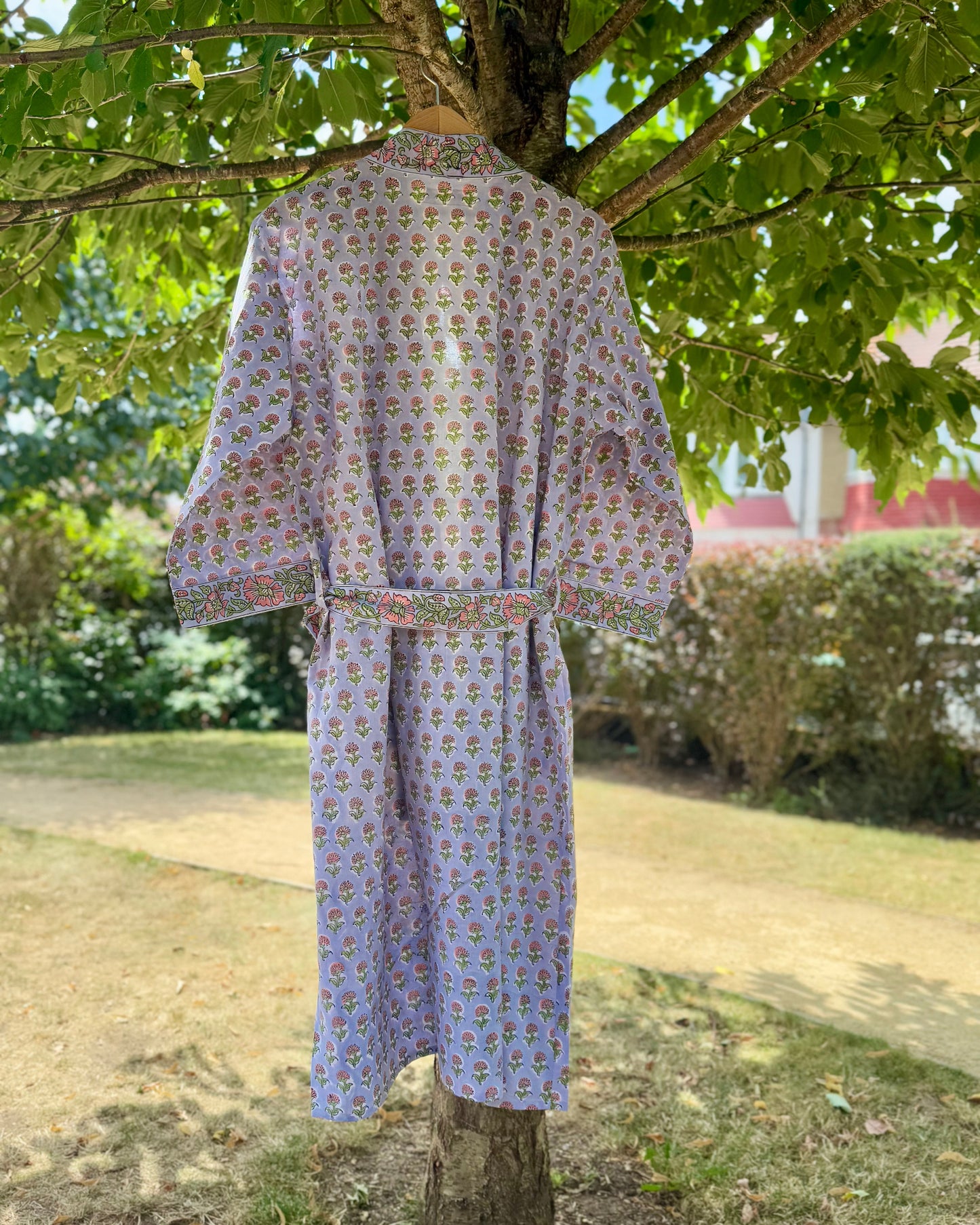 Back view of a floral block printed cotton dressing gown in lilac with pink and green flowers, hanging on a tree in natural sunlight.