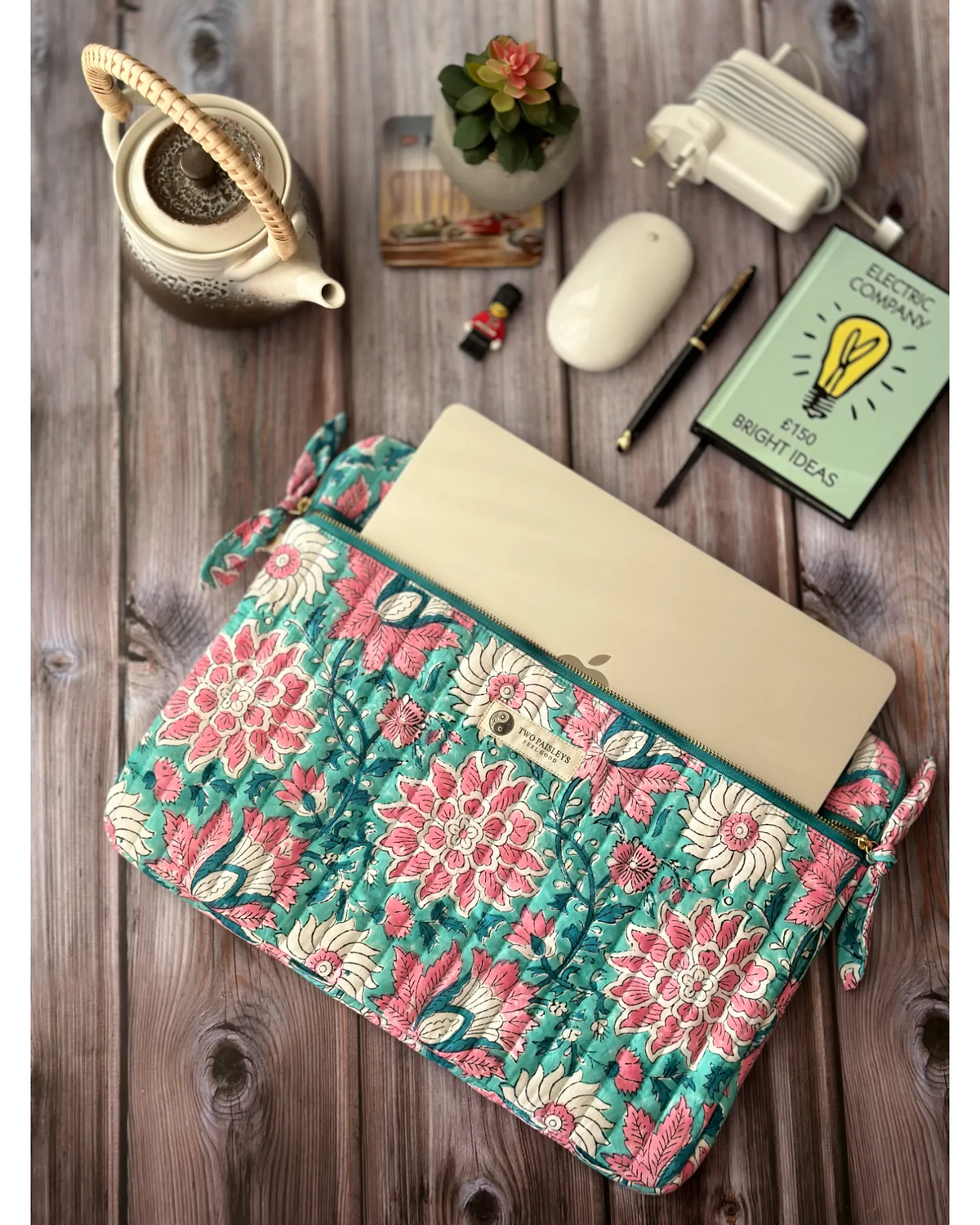 A quilted laptop sleeve with dual zip compartments displayed alongside a 13-inch MacBook Air. Other accessories like a charger, mouse, a notebook and a pen are also visible on the wooden table.