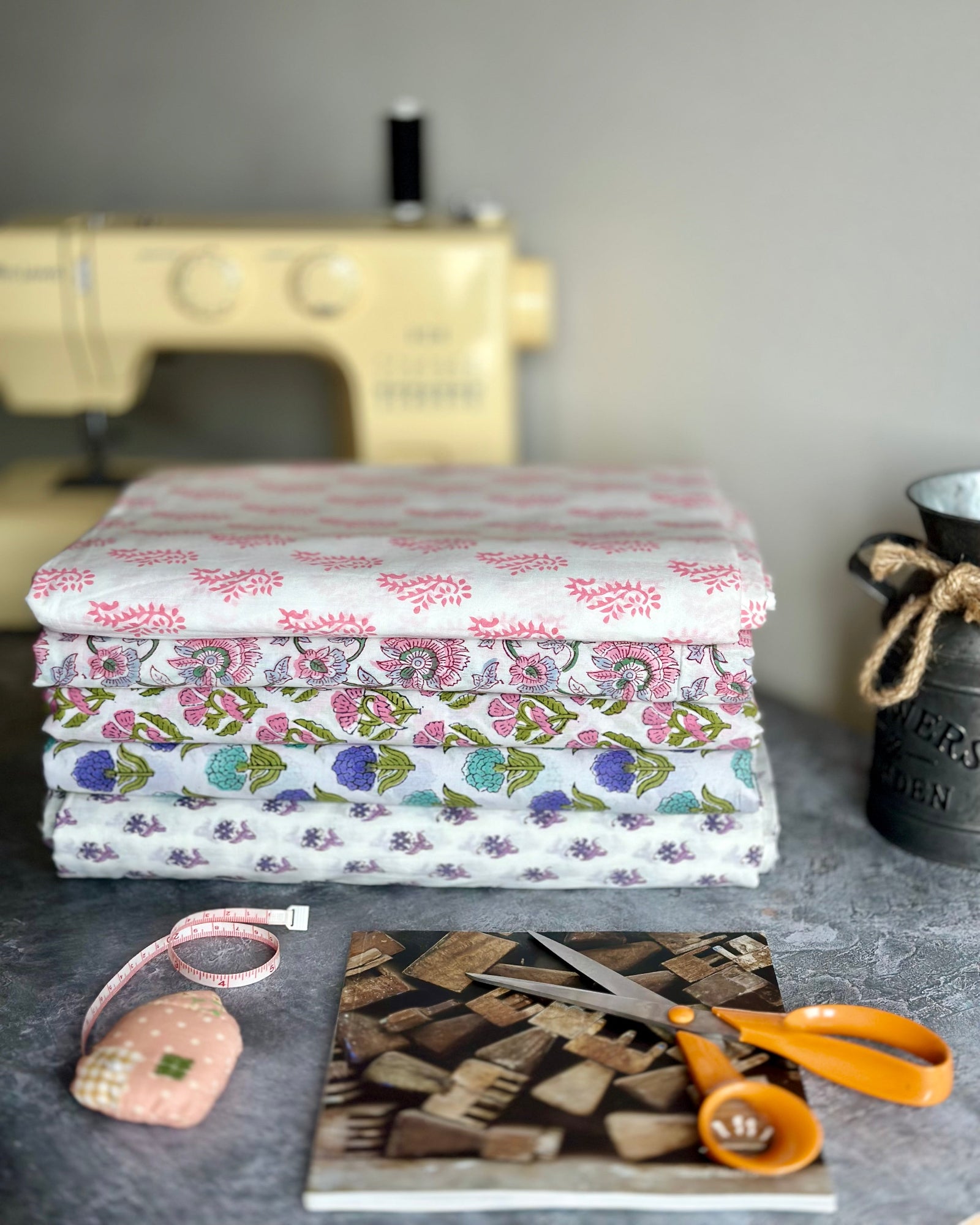 Stack of fabric with a sewing machine, scissors, and measuring tape in the background.