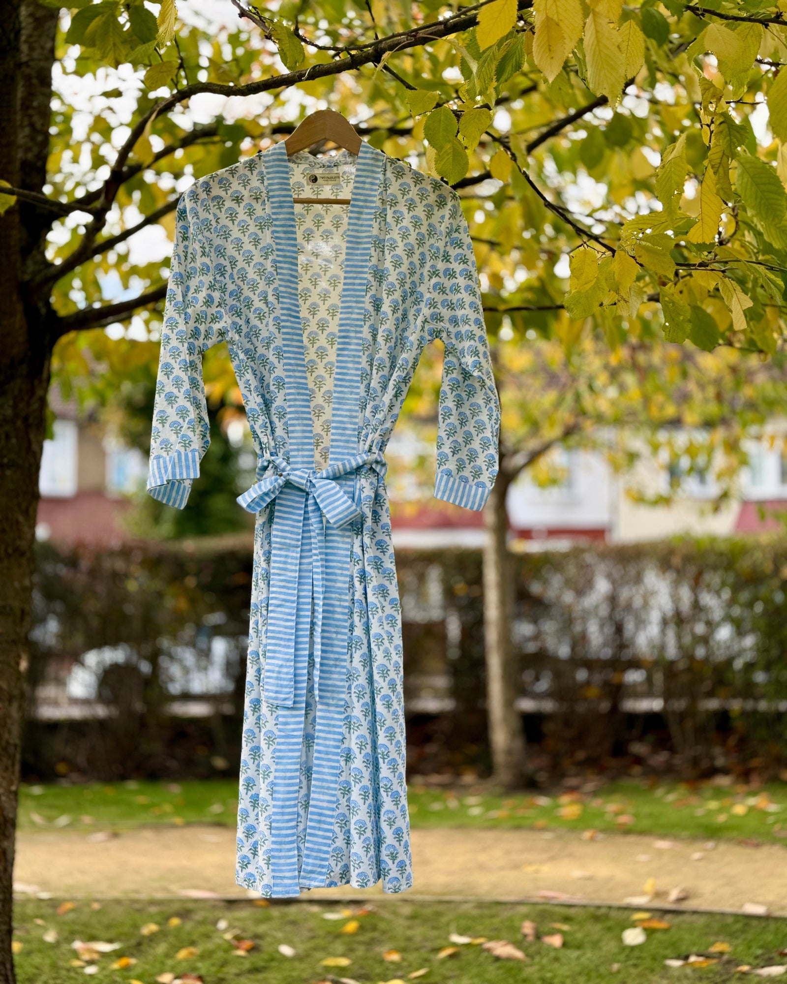A full-length blue blossom hand block print cotton robe with a striped trim and tie belt, hanging from a tree.