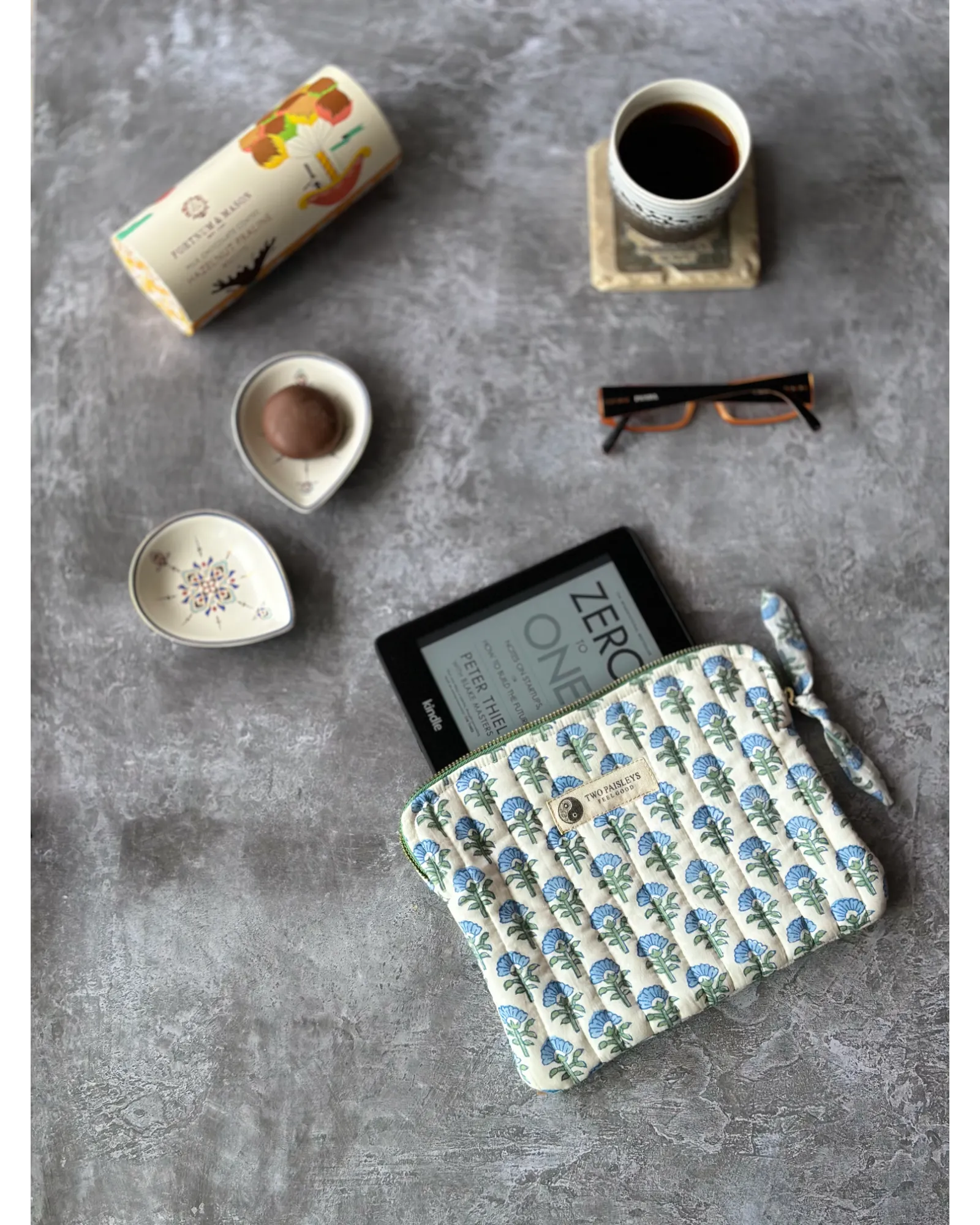 A quilted Kindle cover with a blue floral pattern, displayed on a table with a coffee cup, a bowl, and glasses.
