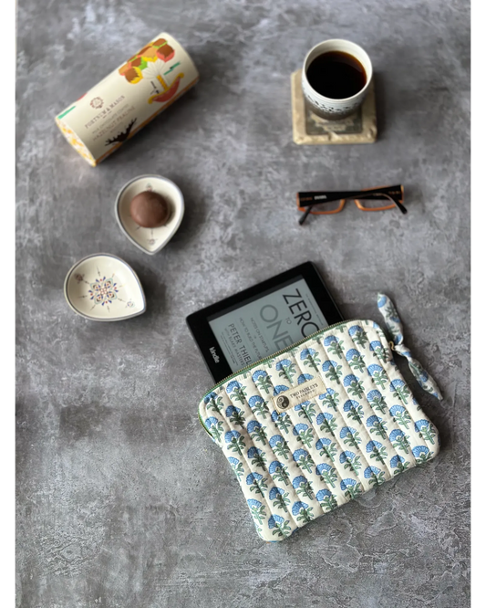 A quilted Kindle cover with a blue floral pattern, displayed on a table with a coffee cup, a bowl, and glasses.