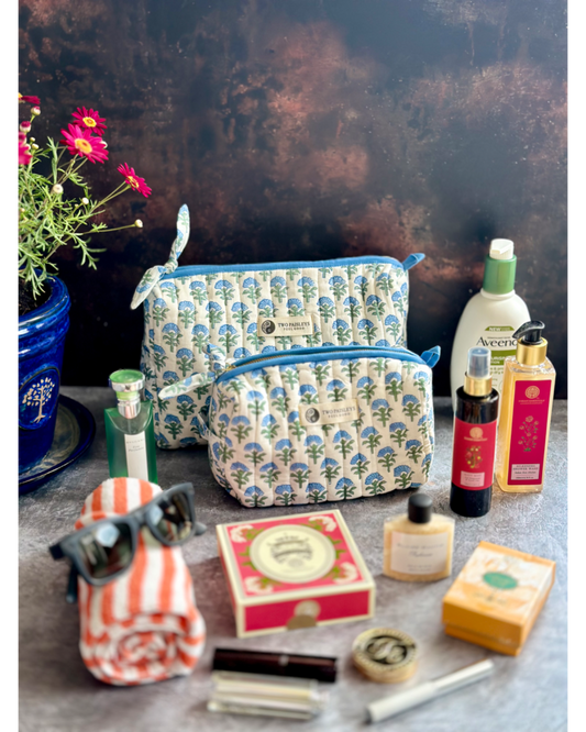 A quilted cosmetic bag with a floral pattern in blue, white, and green shades, displayed with various travel and makeup items like sunglasses, a towel, and bottles.