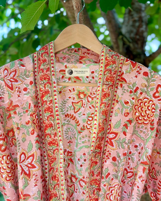 Close-up of a coral pink block printed cotton dressing gown with intricate red and green floral designs and a decorative border, showing the Two Paisleys label.