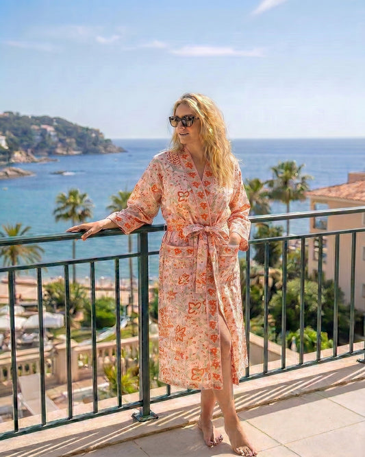 Woman in a floral dressing gown in coral & green hues standing on a balcony with a scenic ocean view.