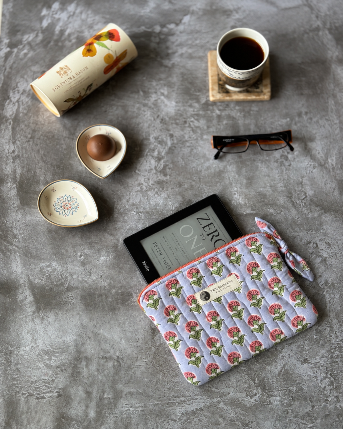 A quilted zip pouch with a pink and green floral pattern on a light lavender background, styled on a gray concrete surface alongside a black Kindle e-reader, a cup of coffee, eyeglasses, and a small dish.