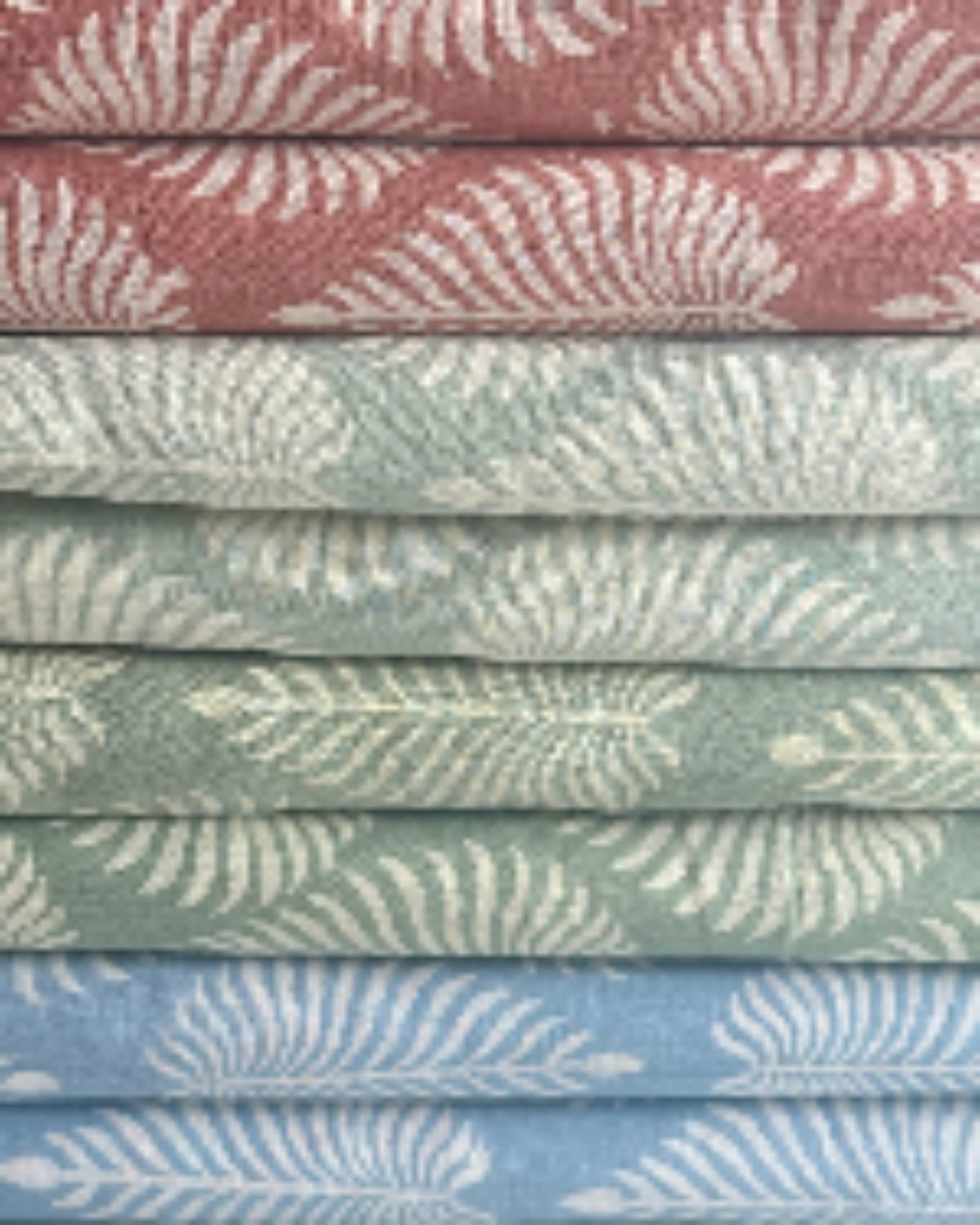 A stack of Fern Garden hand block print cotton fabric in dusty rose, sage green, and sky blue, showcasing the discharge printing technique