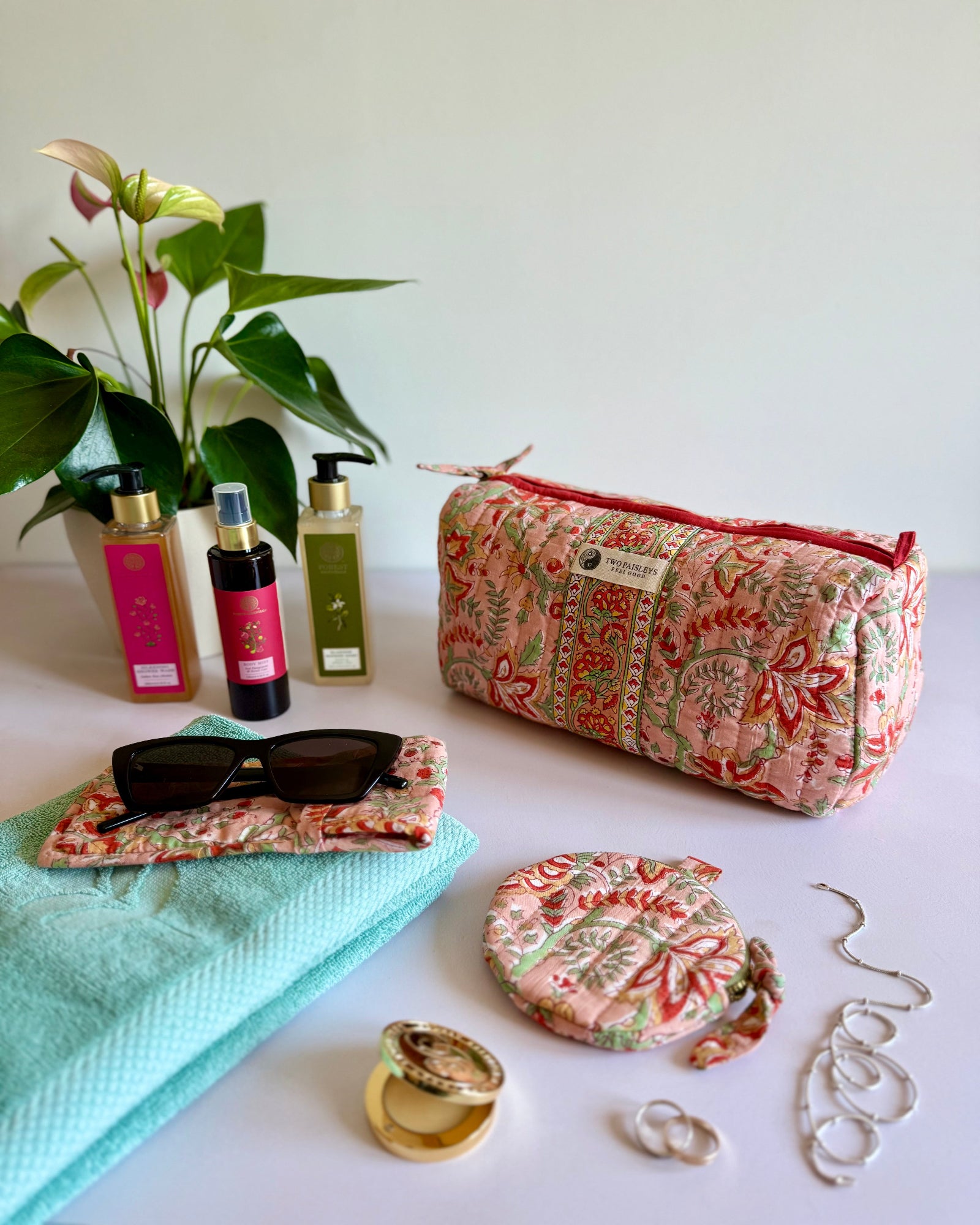Patterned pouch, sunglasses, towel, and jewelry on a white surface with a plant in the background.