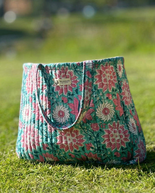 A hand block printed quilted tote bag with a vibrant pink and teal on grass on a warm summer day