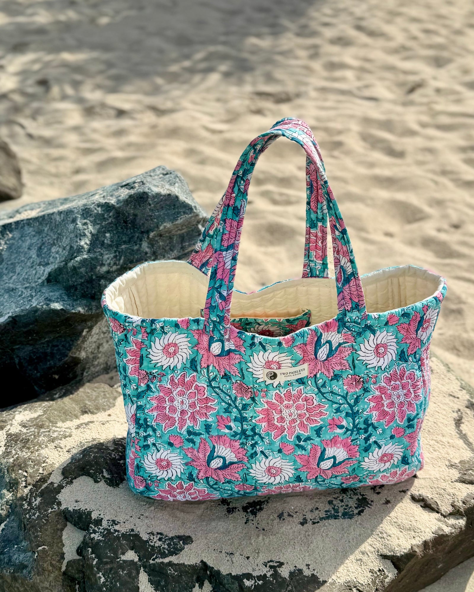 A hand block printed quilted tote bag with a vibrant pink and teal floral design, resting on large rocks on a sandy beach.