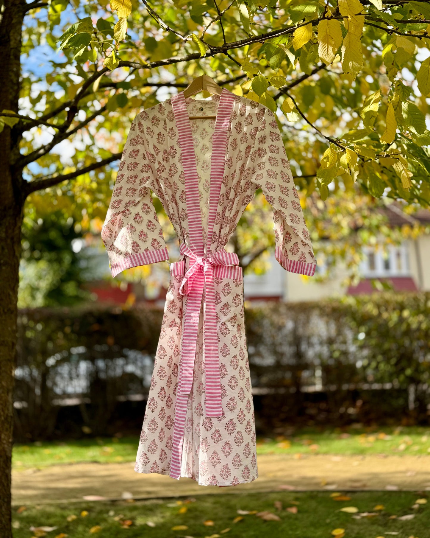 A full-length pink floral hand block print cotton robe with a striped trim and tie belt, hanging from a tree.