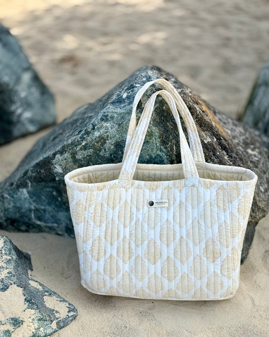 A hand block printed quilted tote bag with an elegant gold and white motif pattern, resting on a large rock on a sandy beach.
