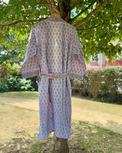 Back view of a floral block printed cotton dressing gown in lilac with pink and green flowers, hanging on a tree in natural sunlight.