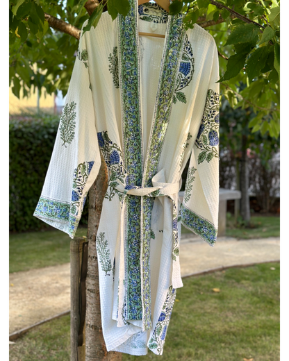 A white waffle cotton bath robe with blue and green floral hand block print, hanging on a branch outdoors.