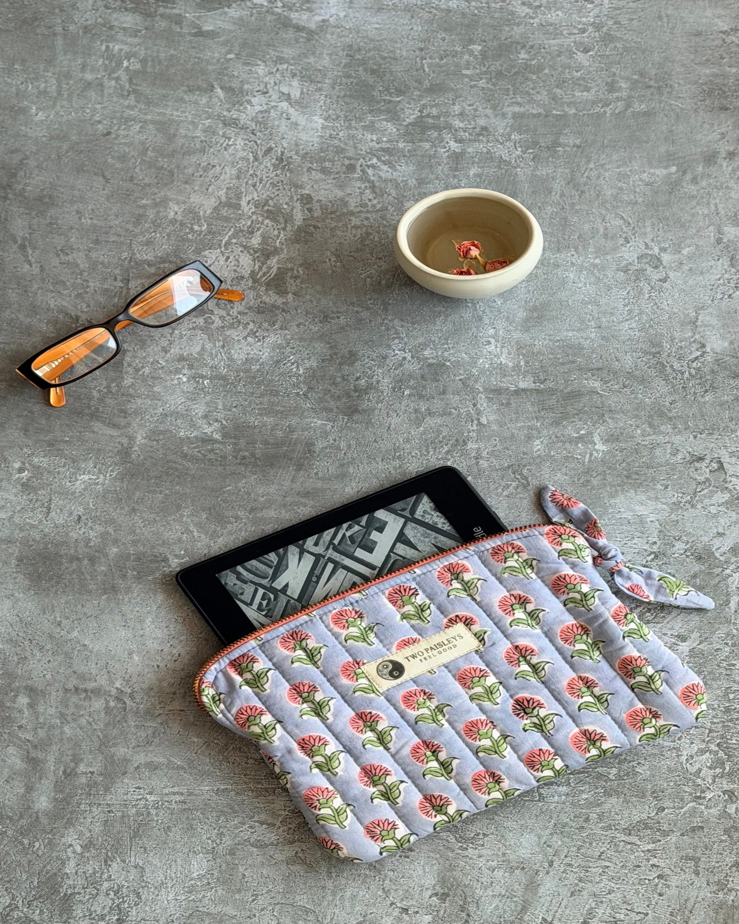Pink florals on a lilac background  patterned pouch, contrast pink zip with a ereader on a grey surface, accompanied by glasses and a cup of  tea