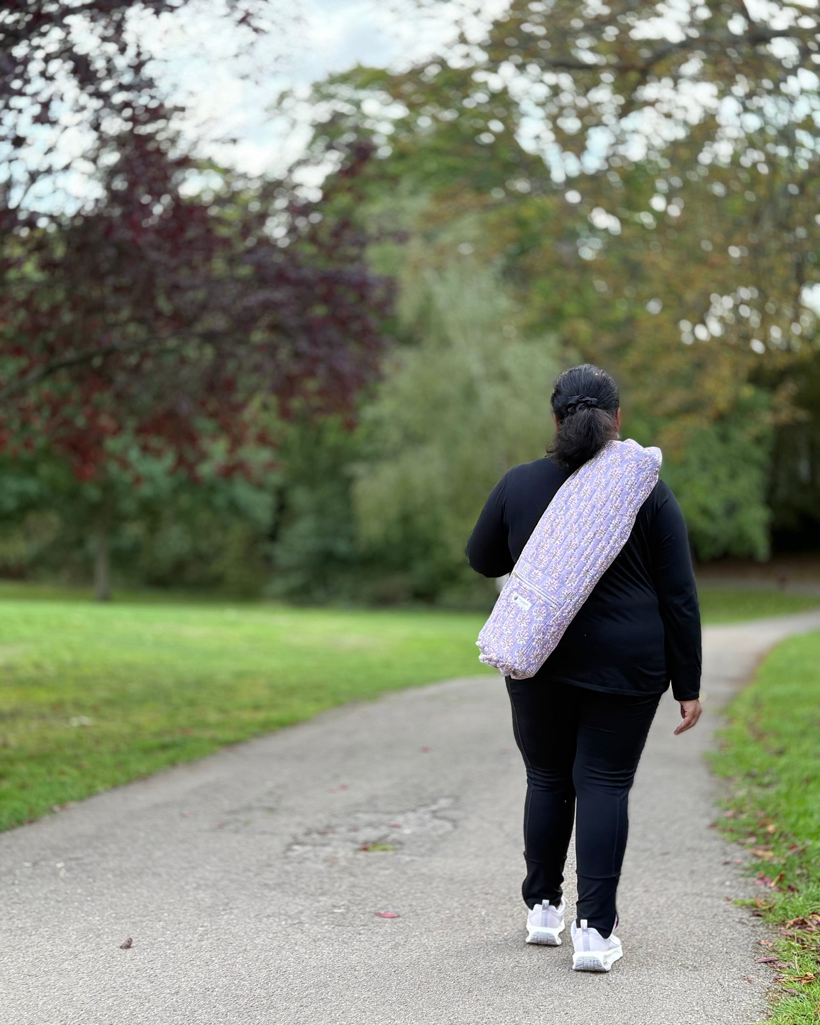 Person walking on a path in a park with a white bag over their shoulder.