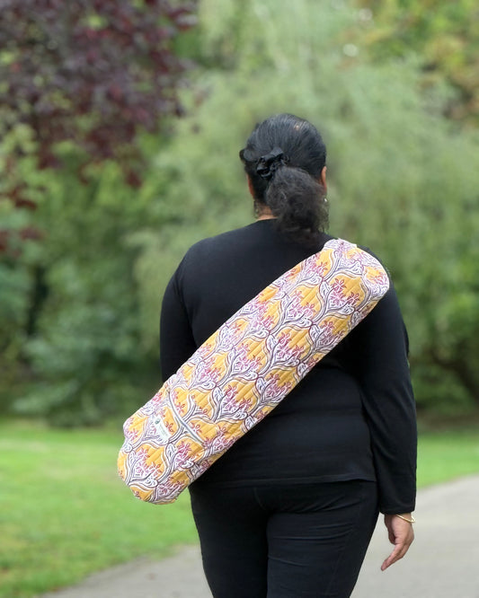 Woman carrying Saffron Song yoga mat bag with golden floral print, crossbody with adjustable strap.
