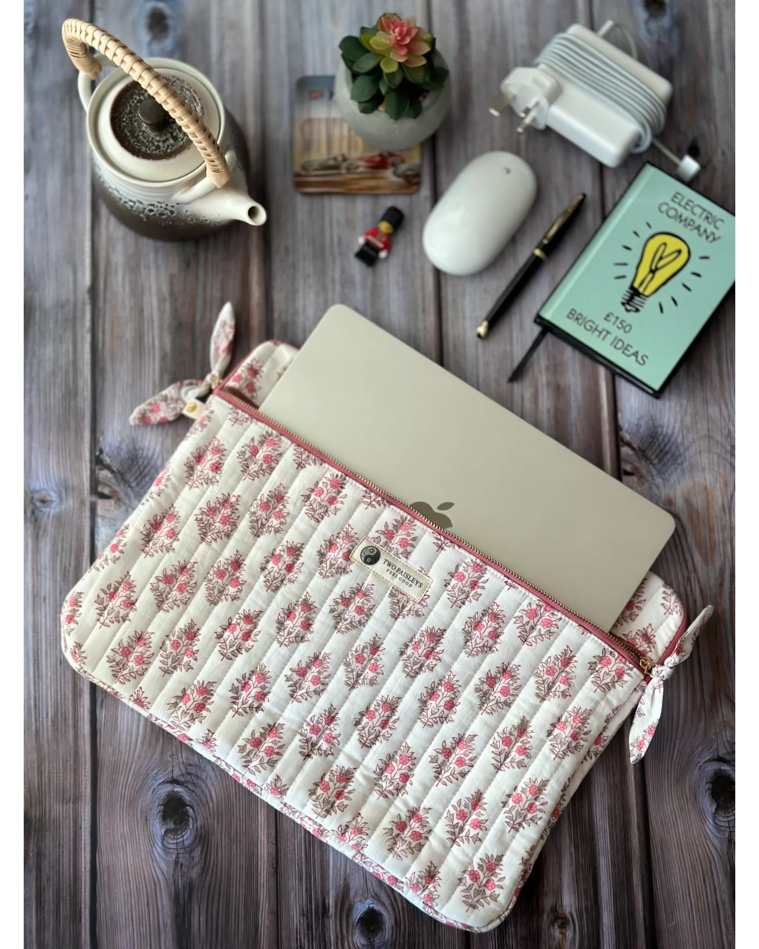A quilted laptop sleeve with dual zip compartments displayed alongside a 13-inch MacBook Air. Other accessories like a charger, mouse, a notebook and a pen are also visible on the wooden table.