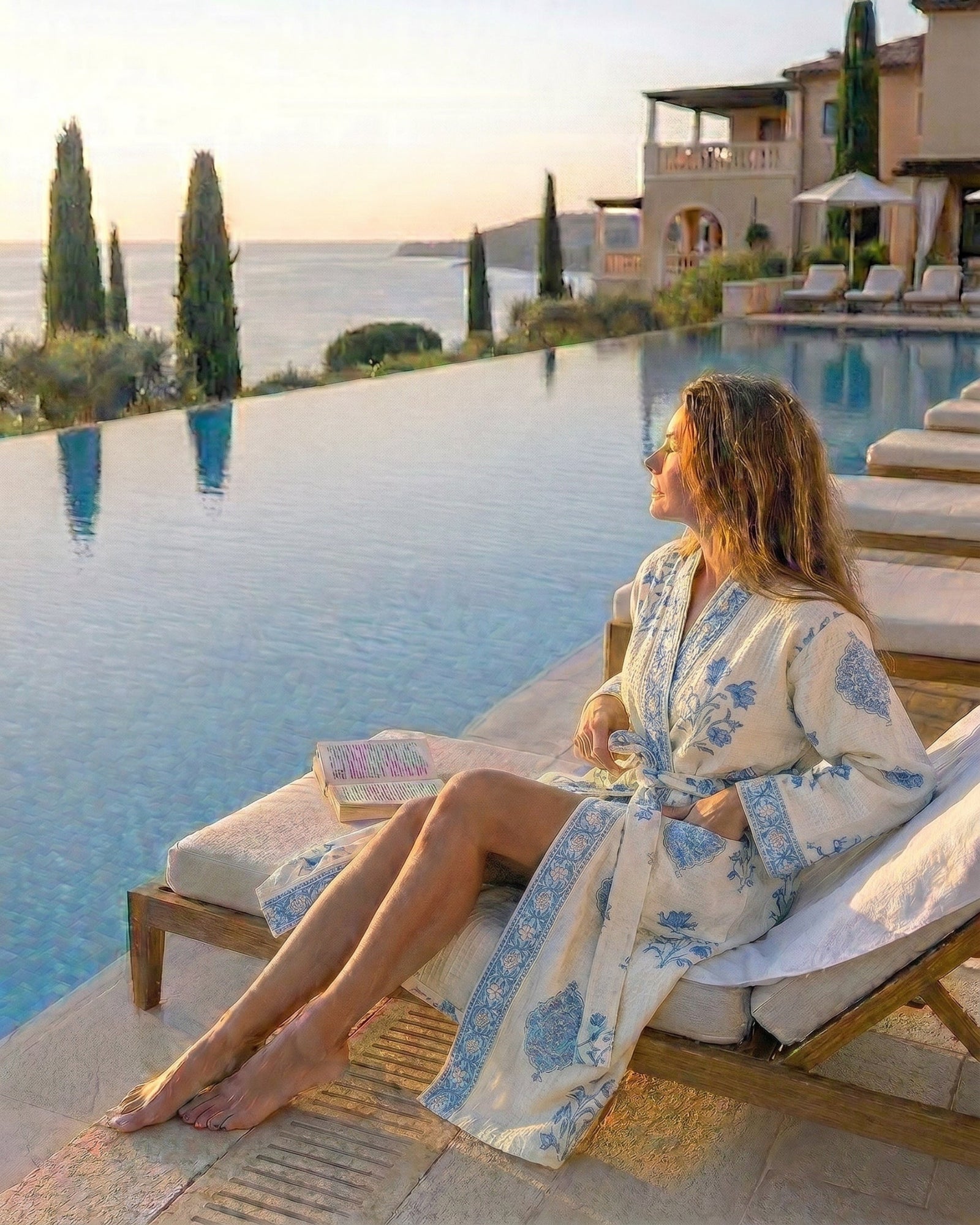 Woman in a waffle robe with light blue motifs on a cream background relaxing by an infinity pool with scenic views.
