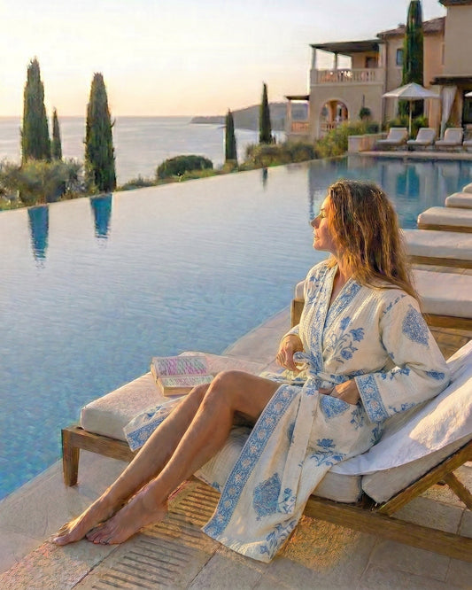 Woman in a waffle robe with light blue motifs on a cream background relaxing by an infinity pool with scenic views.