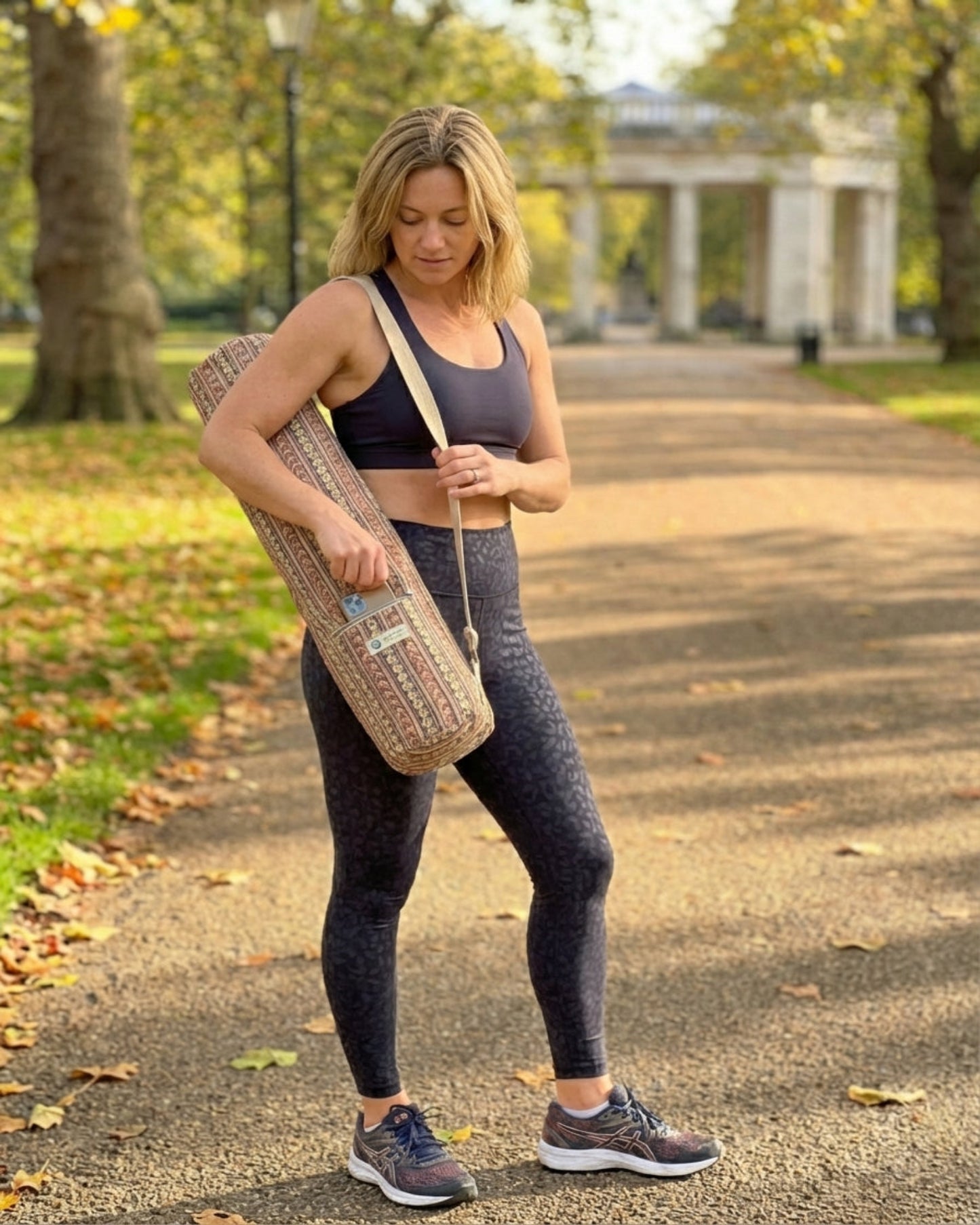 Woman in athletic wear holding a yoga mat bag on a path in a park.