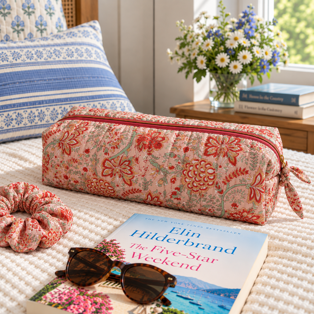 Two Paisleys Coral Floral hand block printed quilted hair tools bag on a bed beside sunglasses and a book for weekend travel