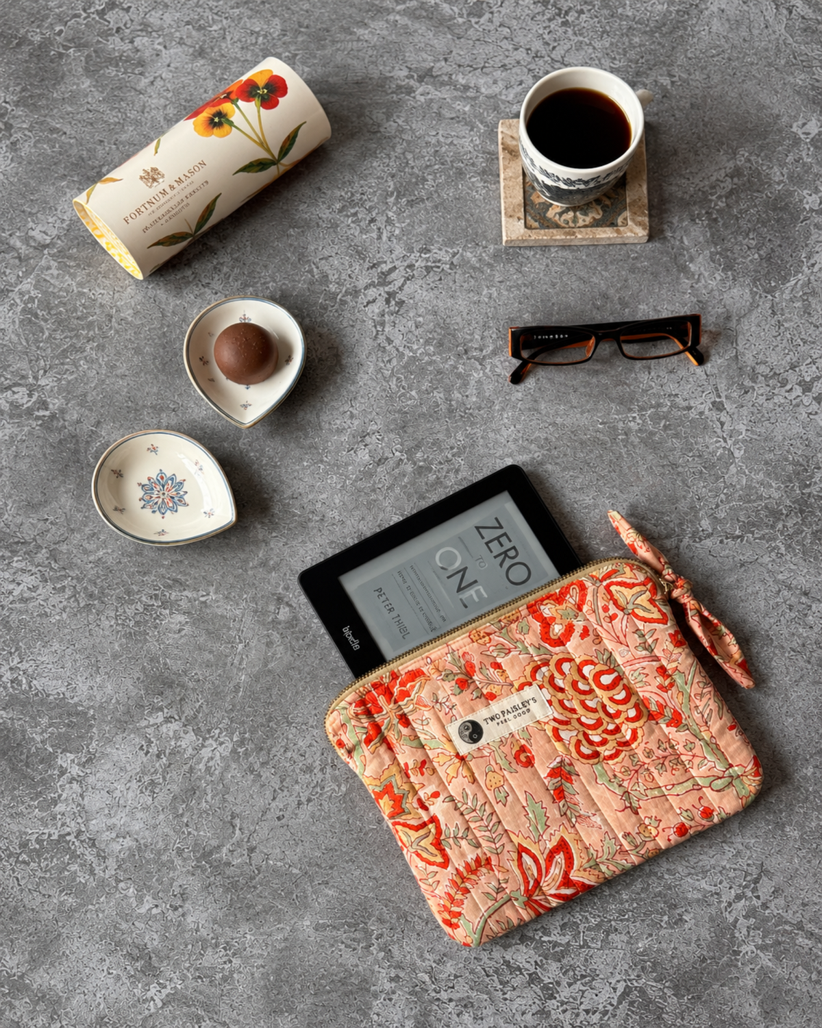 A Quilted Fabric Kindle Sleeve  With A Floral Print In Peach, Orange, And Green, Shown On A Grey Surface Wiith A Black E-reader, A Cup Of Coffee &  Glasses.