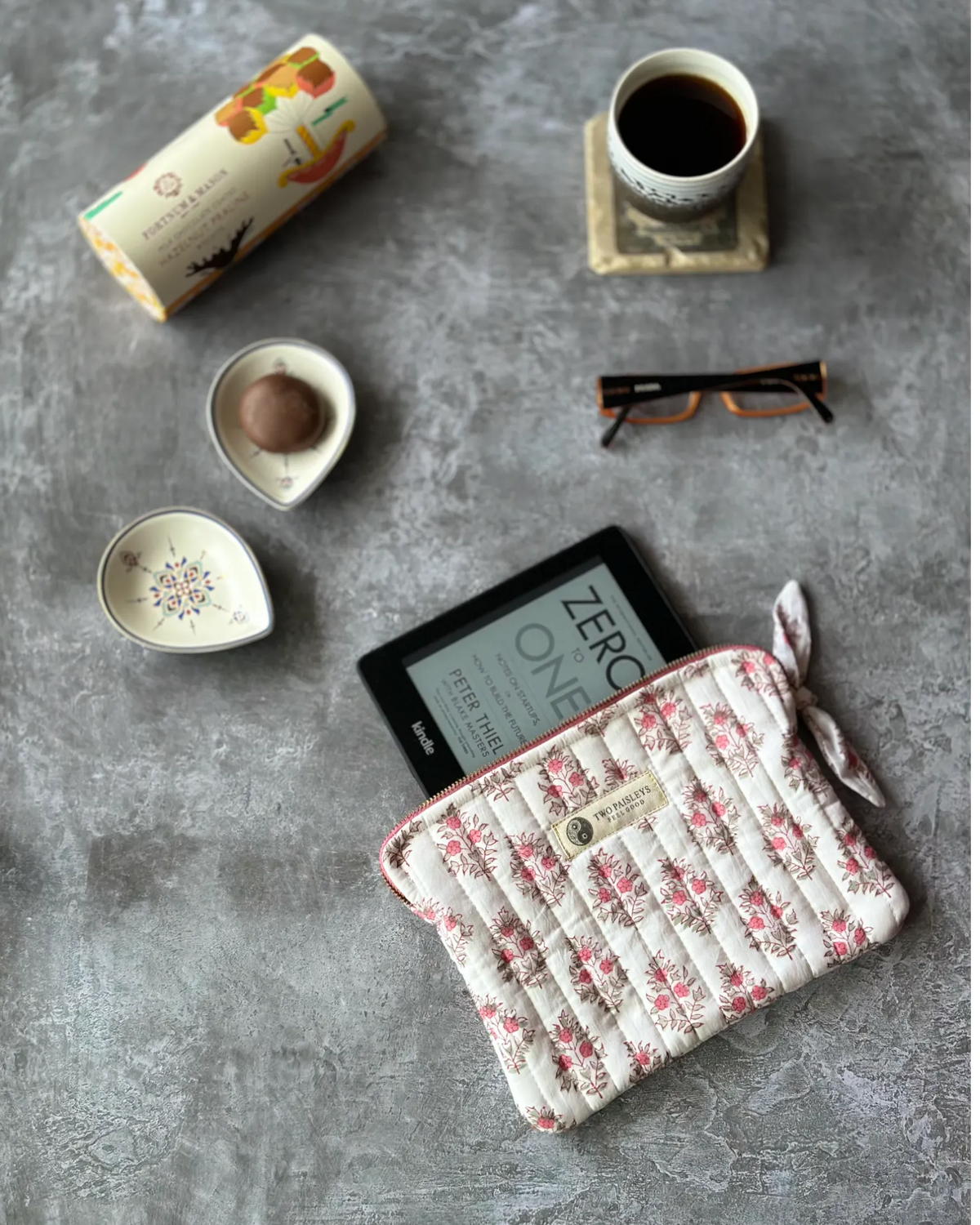 Hand block printed quilted cotton Kindle cover by Two Paisleys in a pink & green floral print along side a cup of tea & some biscuits and reading glasses.
